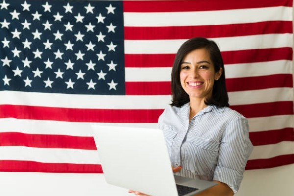 4.visa eb5. una mujer ocn una computadora y al fondo la bandera de estados unidos